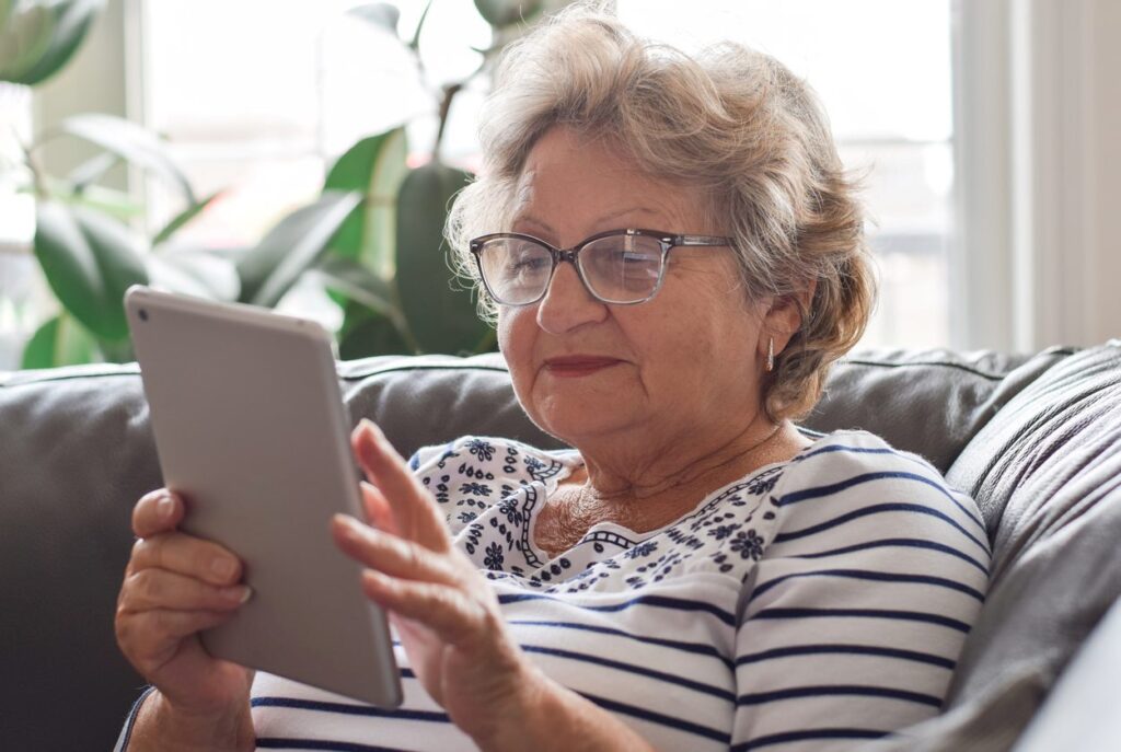 A smiling senior woman in glasses uses a tablet on a comfy couch.