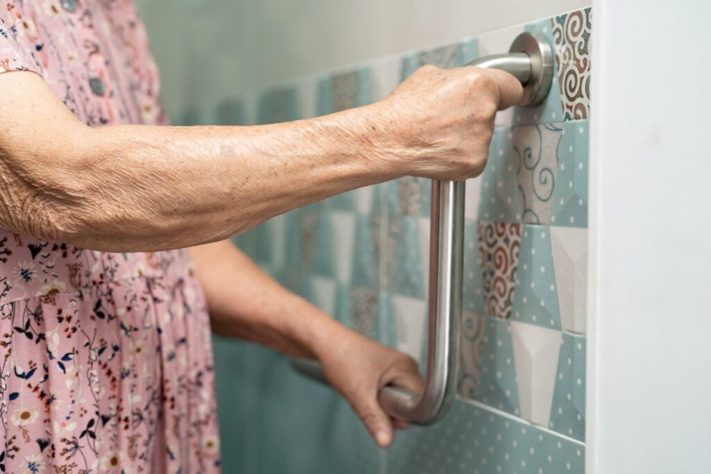 A senior woman in a pink floral shirt uses bathroom grab rails for assistance.