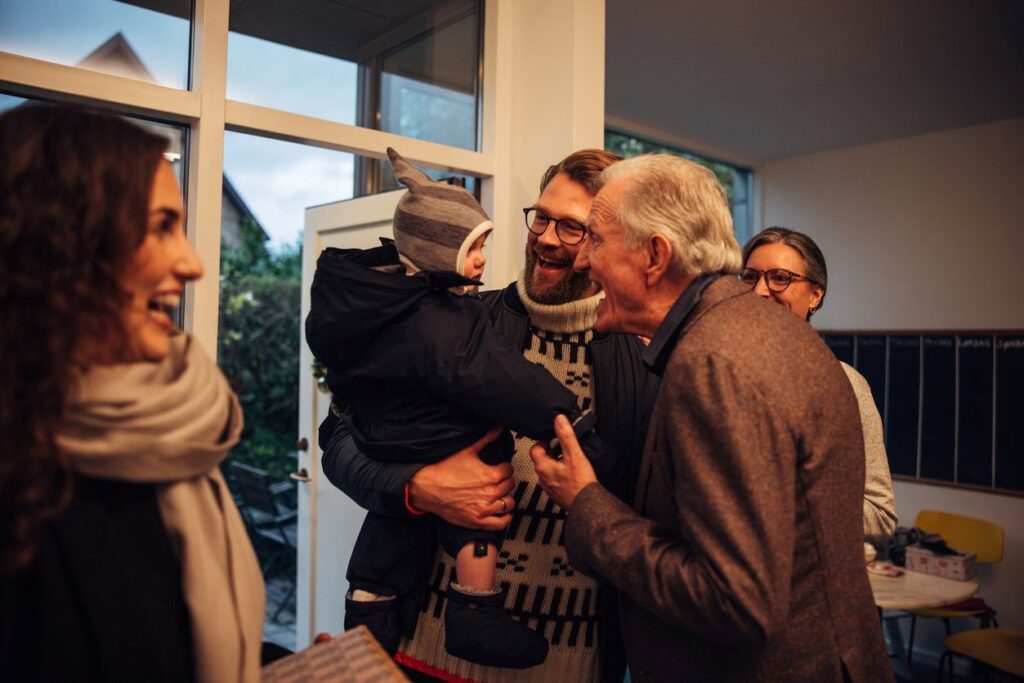 A family arrives at the grandparents’ home for Christmas Eve, with the grandfather smiling at his grandchild.