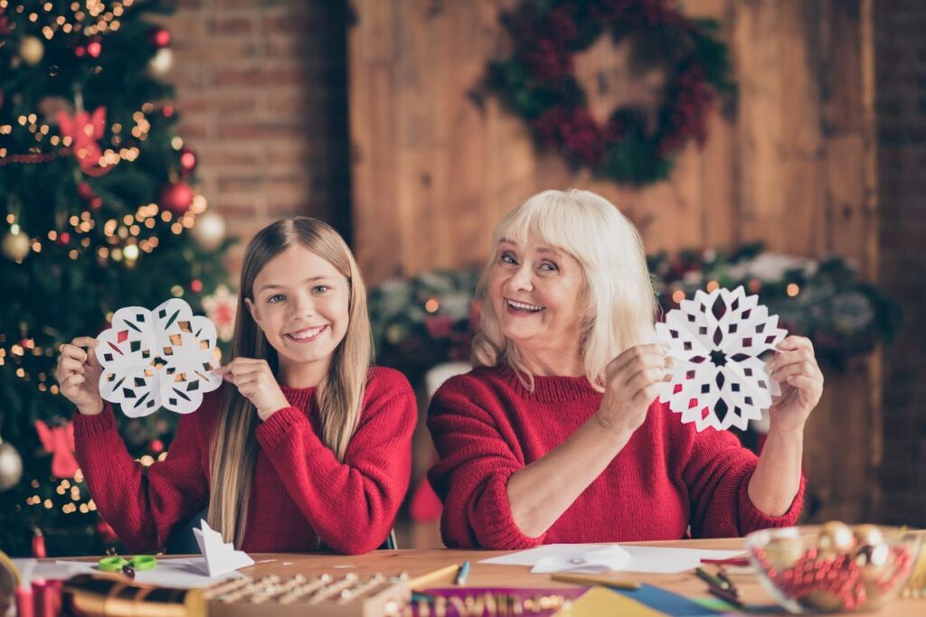 A young girl and her senior woman smile and hold up paper snowflakes during the holiday season.